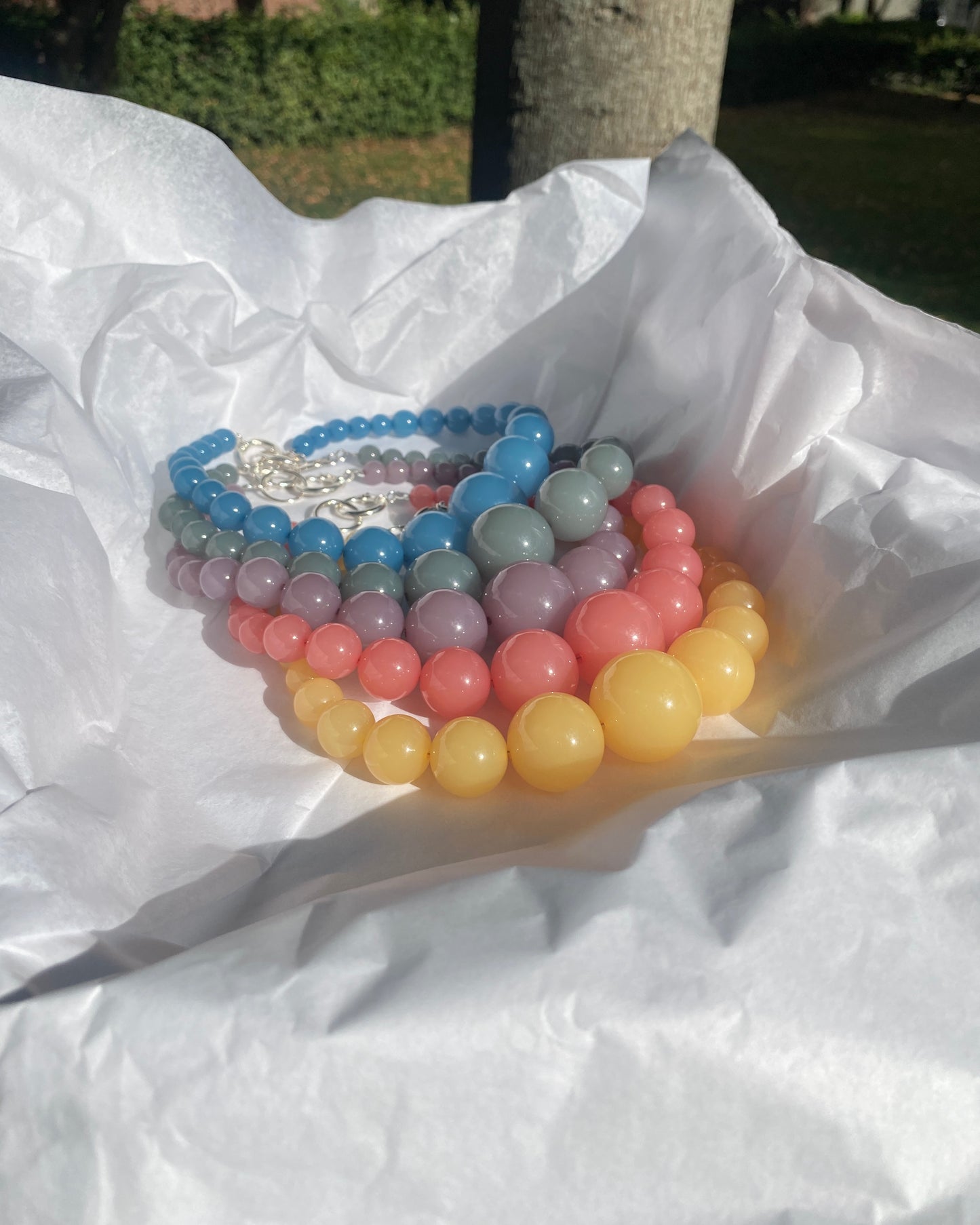 A stack of lemonade beaded necklaces  arranged on crumpled white tissue paper outdoors, with sunlight and blurred greenery in the background.