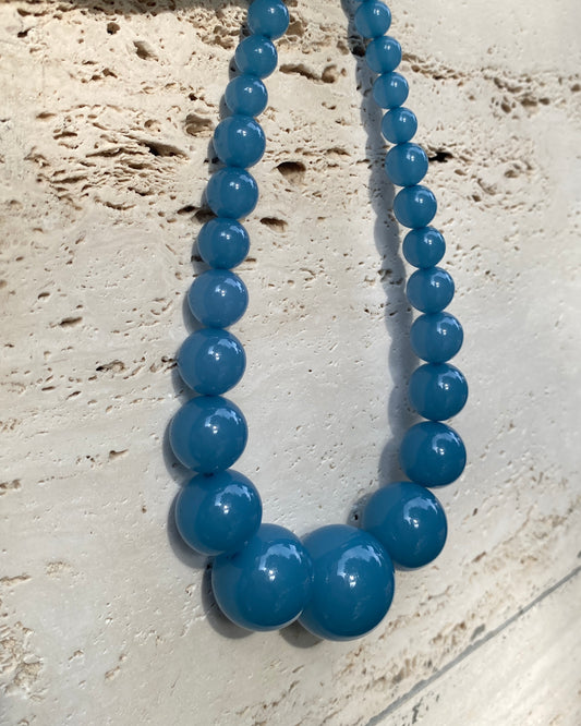 A denim blue beaded necklace with large, graduated glossy round beads on a travertinn wall . made in denmark by lemonade ltd. Daninsk Design
