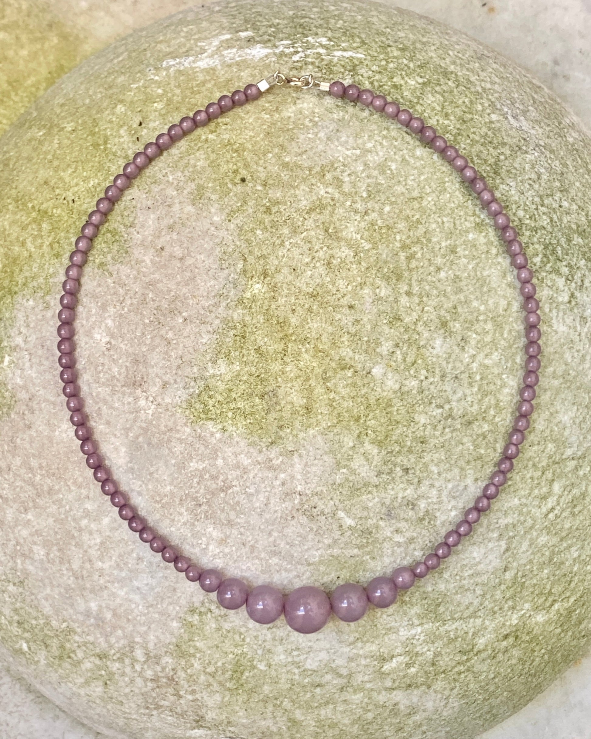 A beaded necklace made of small round lavender-purple beads with three larger matching beads at the front, displayed on a round light green stone. designed by  lemonade denmark