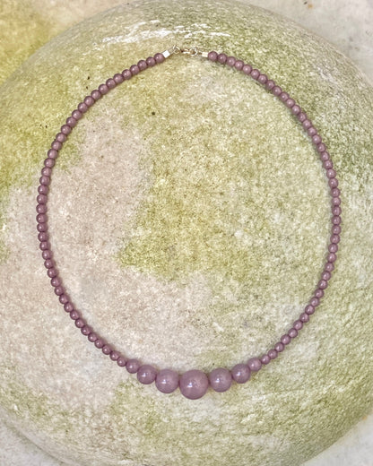 A beaded necklace made of small round lavender-purple beads with three larger matching beads at the front, displayed on a round light green stone. designed by  lemonade denmark