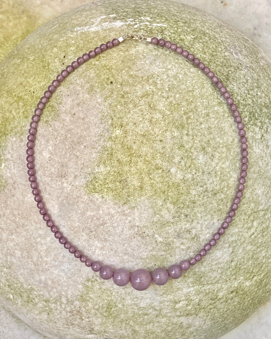 A beaded necklace made of small round lavender-purple beads with three larger matching beads at the front, displayed on a round light green stone. designed by  lemonade denmark